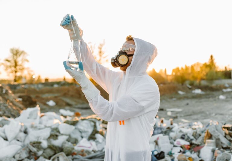 Scientist in protective gear collecting samples at a waste site during sunset.